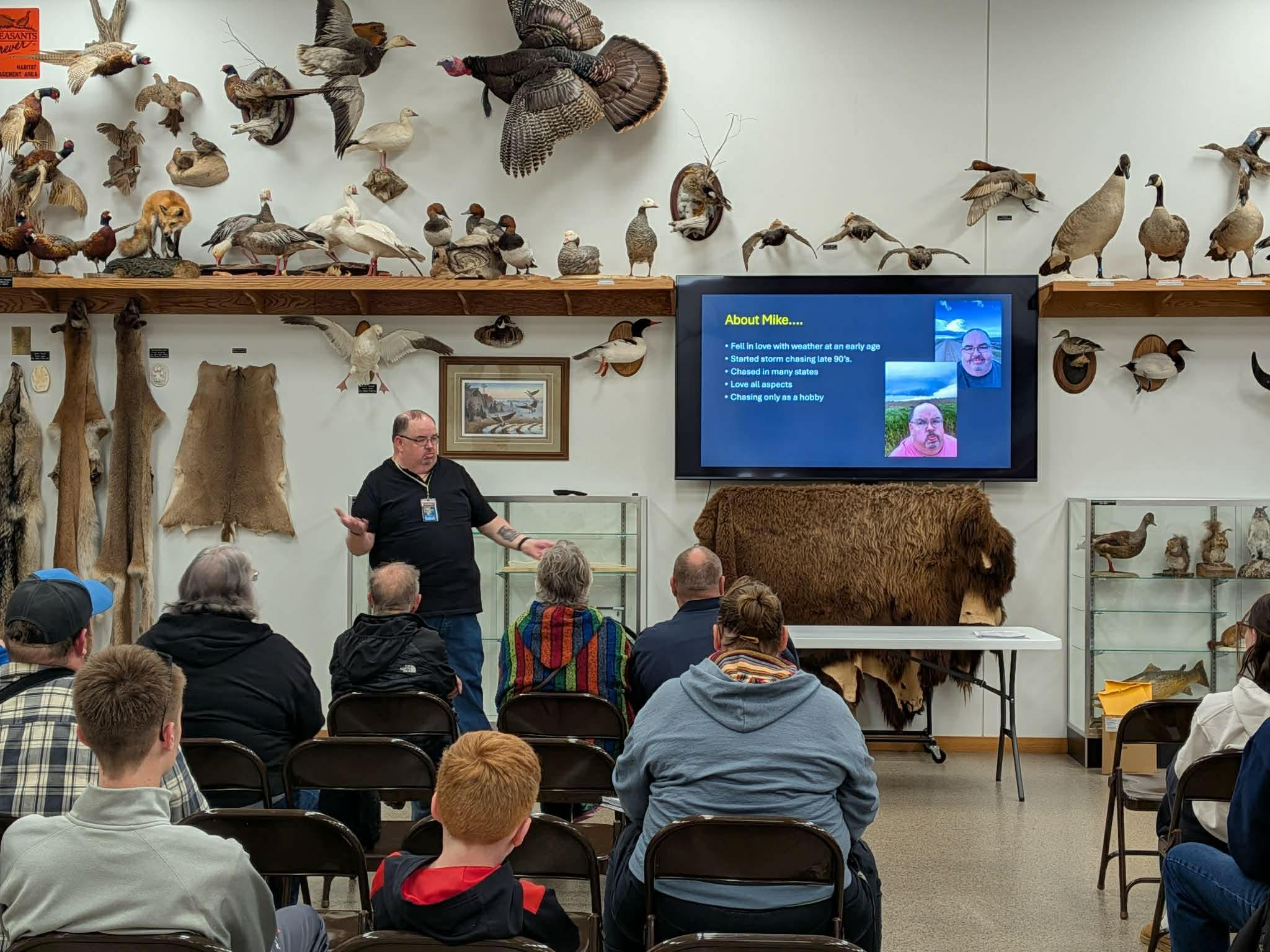 Presenter giving a talk in a museum/taxidermy classroom, with mounted birds and fur displays on the walls and a slide titled 'About Mike...' on a large screen behind him, as audience sits in rows.